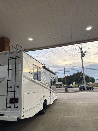 The image depicts a white motorhome parked at a gas station. The scene is captured during daylight hours under a cloudy sky. The motorhome, featuring a ladder at the rear, is positioned near a gas pump under a gas station canopy. Power lines and poles are visible in the background. A dark-colored vehicle is parked beyond the motorhome, along with trees and street lamps. Due to the lack of specific visual identifiers like street signs, or landmarks, pinpointing the exact location within Palm Bay, Florida, is not possible.