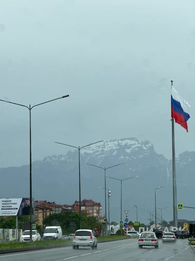 A wide shot captures a city street scene in Oktyabrskoye, Russia, under a cloudy, overcast sky. In the foreground, a wet asphalt road displays lane markings and is traversed by several vehicles. A white sedan with license plate H 761 XK 26 drives away from the camera, followed by a silver sedan. Further back, a white van and a white sedan are also visible. On the left side of the road, a billboard featuring a man's portrait and Russian text reads "ПОДБОР НОВОСТРОЕК МОСКВЫ" (Selection of New Buildings in Moscow), with a phone number. Beyond the billboard, brick buildings and green trees line the street. In the middle ground, a series of streetlights stand tall. In the background, a large, snow-capped mountain range dominates the horizon. To the right, a Russian flag waves prominently from a tall flagpole. Yellow pedestrian crossing signs are visible at intersections. The overall atmosphere is somewhat muted due to the cloudy weather.