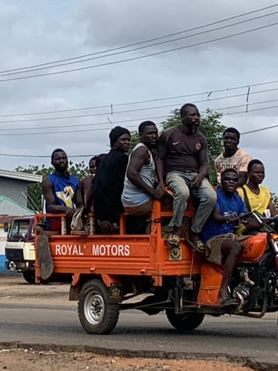 An orange "Royal Motors" branded three-wheeled cargo motorcycle, resembling a motorized tricycle, transports a group of at least nine adult males on a paved road in Shendam, Nigeria. The vehicle is in motion under an overcast sky, with multiple power lines visible horizontally in the upper frame.
Five males are positioned within the open cargo bed. One male on the left wears a blue sleeveless shirt with a white and yellow design. Another male in the center-left wears a black long-sleeved shirt and a black beanie. A third male in the center wears a light blue sleeveless top. A fourth male with a beard, wearing a dark brown short-sleeved shirt and light-colored jeans, sits on the side rail of the cargo bed. A fifth male wears a light-colored "ashlux" t-shirt. Two additional males are partially visible within the cargo bed, behind the first group.
Two males occupy the front motorcycle section. The driver, wearing a blue short-sleeved t-shirt and dark sunglasses, operates the handlebars. A passenger in a yellow t-shirt sits directly in front of the driver, facing forward.
The vehicle displays "ROYAL MOTORS" in white lettering on its orange cargo bed. It features knobby tires and a crowned logo near the rear wheel. A dark-colored mud flap hangs from the left side of the cargo bed. A light-colored container is secured to the frame near the driver's feet.
In the background, a light-colored truck is visible to the left, and a building with a blue-green roof and white wall is further behind it. Green vegetation is present behind the vehicle. The road surface is asphalt, with a visible shoulder or median strip to the right.