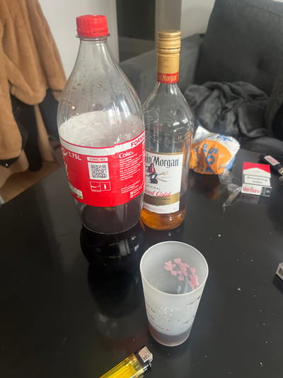 This image depicts an indoor scene on a dark, reflective table surface, likely within a private residence in Saint-Just-en-Chaussée, France. Prominently displayed are a partially consumed 1.75L plastic Coca-Cola bottle with a red label and a nearly empty glass bottle of Captain Morgan Original Spiced Gold rum, featuring a gold cap. A frosted glass cup, partially filled with a dark liquid and containing a small pink decorative floral element, rests nearby. Also on the table are a red and white pack of Marlboro cigarettes bearing the warning "Smoking kills," a yellow translucent disposable lighter, and a second black and silver lighter. In the background, a dark gray fabric sofa is partially visible with a dark blanket draped over it, along with a crumpled white and orange plastic bag. To the far left, a light brown furry garment or blanket is present. No individuals are visible.