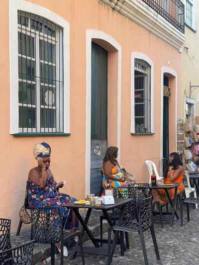 Three women sit at tables outside a building with peach-colored walls. The women are wearing colorful outfits. Two tables have drinks and other items on them. The building has windows with security bars and arch-shaped tops. A few items are hanging on the building wall behind one of the women. The area appears to be a restaurant patio.