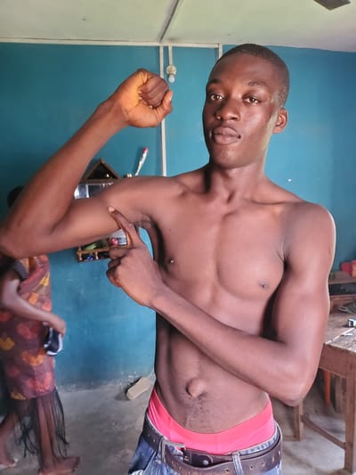 A shirtless dark-skinned man is indoors. He is wearing jeans with a brown belt and red underwear showing at the waist. He is flexing his left arm and pointing to his right arm. Another person is lying on the floor in the lower portion of the image, possibly asleep and covered. The location is Atani, Nigeria. The room has turquoise walls.