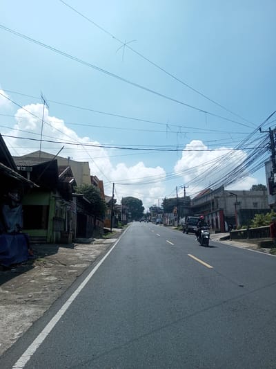 The image depicts a street in an urban area. The road is asphalt with a single yellow dividing line and white edge lines. A person on a moped is traveling towards the camera. Buildings of varying styles and ages line both sides of the street. An extensive network of overhead wires spans the street, partially obscuring a blue sky with scattered clouds. There is no specific evidence to confirm that this is located in Zeyathiri District, Myanmar.