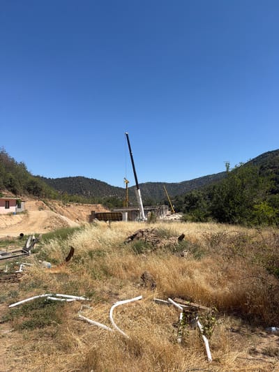 An outdoor, wide-angle shot captures a construction site on a bright, clear day, set against a backdrop of rolling, tree-covered hills. In the foreground, dry, golden grass and various construction materials, including white pipes and metal framing, are scattered across the uneven terrain. Further into the scene, a dirt road winds up a dusty hillside towards a small, pink building on the left. The central focus is a partially constructed bridge or structure, with scaffolding and concrete forms visible. Two cranes are present: a large white crane on the right and a smaller yellow crane in the middle distance. The overall impression is one of an active, ongoing construction project in a somewhat arid, natural environment.