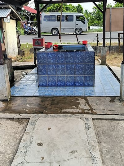 This outdoor scene, likely in or near Blangbitra, Indonesia, features a tiled ablution facility under a simple open structure. The facility is rectangular, constructed with blue tiles bearing a leaf pattern, and contains water. Red containers, possibly for scooping water, sit on top. The surrounding area has light blue tiled flooring around the ablution basin and sections of exposed concrete that are stained and show signs of weathering.
In the background, a white minivan travels on an adjacent road, and two individuals are seen riding on a scooter on the opposite side of the road. Beyond the road are trees and other vegetation. The scene is captured in daylight.