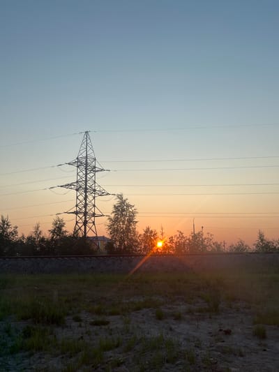 This is a landscape photograph taken in Novy Urengoy, Russia, oriented vertically. The scene depicts a sunset with a soft gradient of blue transitioning to a warm orange glow in the sky. Several power lines are visible, stretching horizontally across the frame. In the center, a tall, lattice-style transmission tower is silhouetted against the sky.
To the right of the tower, trees with sparse foliage are present, and the setting sun is a bright, distinct orb peeking through the branches, casting a lens flare. Further to the right, a rough, unpaved ground surface with scattered grass and some debris is visible, suggesting an outdoor, possibly rural or undeveloped area. A railway track runs along the edge of this ground. In the background, beyond the trees and power lines, a building with an orange roof and yellow walls is partially visible. The overall mood is serene and tranquil, characteristic of a quiet evening. The time of day is clearly sunset, indicated by the warm, low-angled sunlight and the colors in the sky. There are no people visible in the image, and no text is present.