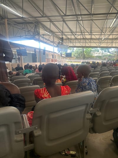 The image depicts an indoor scene within a large, open-sided structure, likely a hall or event space, in Ozuaha, Nigeria. The perspective is from behind several rows of seated individuals, looking towards a stage area.
In the immediate foreground, two young children are visible from the back, seated in grey, foldable stadium-style chairs. The child on the left wears a red dress with a gold patterned design. The child on the right, appearing younger, wears a blue and white patterned garment and has a gold hoop earring in their visible left ear; this child is slightly turned to their left.
Behind these children, numerous individuals are seated in uniform grey chairs, extending in multiple rows towards the background. Most are viewed from behind, their attention directed forward.
In the mid-ground, an elevated stage area is visible. A person stands at a podium on the stage, addressing the assembly. Two large screens, one on each side of the stage, display visual content. Behind the stage, a light-colored building structure with brown railings is present.
The overhead structure features a metal truss roof with exposed beams and fluorescent strip lighting fixtures. The open sides of the building allow significant natural light to enter and reveal tropical foliage and the sky in the distant background. The floor appears to be concrete or paved. The overall scene suggests a public gathering, possibly a religious service or community event.