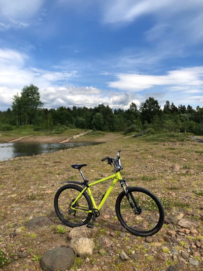 A bright yellow mountain bike is parked on a rocky, grassy embankment overlooking a body of water. The bike is positioned in the foreground, with its front wheel pointed slightly to the right. Behind the bike, a calm body of water reflects the blue sky and scattered white clouds. The far bank of the water is lined with a dense forest of green trees. The ground where the bike is parked is covered with stones of various sizes and patches of dry grass, with some small pink and yellow wildflowers scattered throughout. The overall scene appears to be a natural, outdoor setting, possibly a recreational area near Svirstroy, Russia, given the environment. No people are visible in the image.