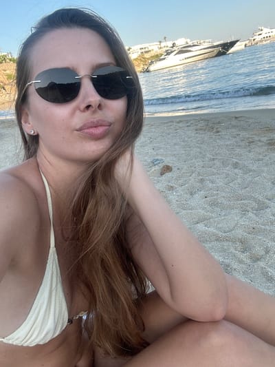 A young woman with long, brown hair and sunglasses is sitting on a sandy beach in Psarou, Greece. She is wearing a light-colored bikini top and is leaning on her hand, with her legs crossed. In the background, the blue sea is visible with several yachts anchored in the water. The sky is clear and blue, indicating a sunny day.