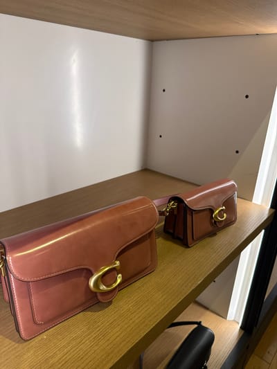 The image shows a display of brown leather purses with gold hardware in a retail setting. The purses are stacked on wooden shelves illuminated by strip lighting. The location is likely within the city of Sunrise, United States of America.