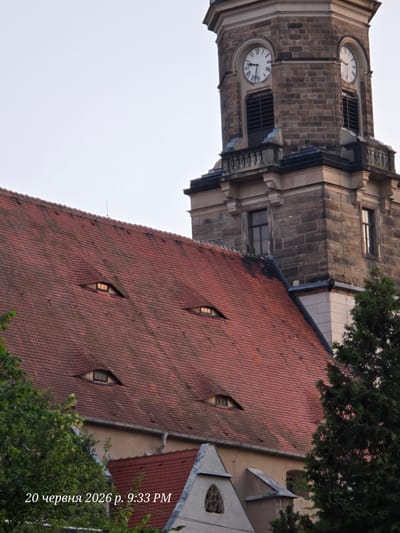 The image shows a close-up view of a church tower and a red-tiled roof with several dormer windows that resemble eyes. The church tower, made of stone blocks, features two clock faces with Roman numerals. The left clock shows approximately 7:40, and the right clock shows approximately 9:05. The roof has a distinctive eye-like shape due to the dormer windows, giving the building a watchful appearance. Trees and vegetation are visible in the foreground and to the right of the tower. The sky is overcast. The text "20 червня 2026 р. 9:33 PM" is superimposed on the bottom left of the image. The location of Bannewitz, Germany, is mentioned, suggesting this is likely the historic church in Bannewitz.