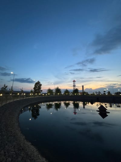 The image depicts a tranquil dusk scene at a retention pond in Novaya Adygeya, Russia. The sky transitions from a deep blue overhead to lighter hues near the horizon, with scattered clouds reflecting the fading daylight. Along the curved perimeter of the pond, a gravel embankment is visible, topped by a railing with numerous small, illuminated lights. Trees and industrial structures, including tall towers with illuminated tops, are reflected in the still water. A small, dark, roofed structure is positioned in the pond, its reflection also visible. The overall atmosphere is serene, suggesting a public park or recreational area.