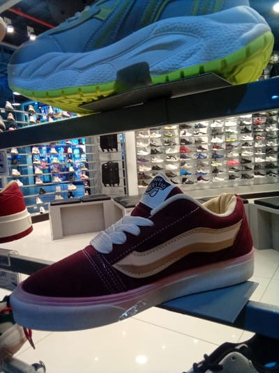 The image captures a display inside a shoe store in Parroquia Guatire, Venezuela. Numerous pairs of shoes are neatly arranged on shelves lining the walls. In the foreground, a maroon and tan Vans shoe is prominently showcased on a display shelf. Above it, another shoe, bluish in color with a yellow sole, sits on a higher shelf. The store is brightly lit, and the floor is white tile.