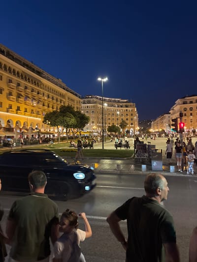 This is an outdoor, eye-level shot of Aristotelous Square in Thessaloniki, Greece, at dusk or early evening. The sky is a deep blue, indicating the transition from day to night. The square is illuminated by streetlights and the warm glow emanating from the buildings.

In the foreground, three people are visible. On the left, a man wearing a dark green polo shirt is seen from the back, looking towards the left. Next to him, a young girl with brown hair and sunglasses on her head is in focus. She is wearing a light-colored top and appears to be looking at the man, with her right arm raised slightly. To the right of the girl, a man with thinning grey hair and a dark green polo shirt is seen from the side, looking to the right.

A dark-colored car is in motion on the wet street in the mid-ground, its headlights illuminated, creating streaks of light due to motion blur. Beyond the car, the square unfolds with a lush green lawn area dotted with trees. People are scattered throughout the square, some sitting on benches, others walking, and many dining at outdoor cafes under umbrellas.

The architecture of the surrounding buildings is distinctive, featuring multi-story structures with arched windows and balconies. The buildings are illuminated, casting a warm light that contrasts with the darkening sky. In the background, the street extends into the distance, lined with more buildings and activity. A traffic light is visible on the right side of the frame, showing a red signal. Graffiti is present on a structure in the mid-ground. The overall atmosphere suggests a vibrant urban evening scene with people enjoying the public space.