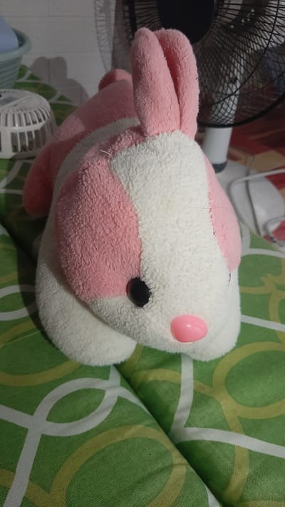 A plush toy bunny, predominantly white with pink accents and stripes, lies on a green and white patterned surface. The bunny has pink floppy ears, a black eye, and a pink nose. In the background, a black electric fan is visible, along with a white ribbed object and a glimpse of a blue bowl. The scene appears to be indoors, possibly a bedroom, in Kawit, Philippines.