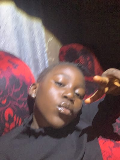 The image is a blurred, close-up selfie of a person with dark skin, appearing to be a young individual, possibly female, due to the earring visible. They are wearing a dark-colored top, and their face is illuminated by a bright light source, creating highlights on their skin and lips. The background is indistinct but appears to feature a pattern of red and black shapes against a lighter, possibly textured surface. The overall impression is of an indoor setting, though the specifics are obscured by the blur and low light. There is no identifiable information to suggest a location in Russo, Nigeria.