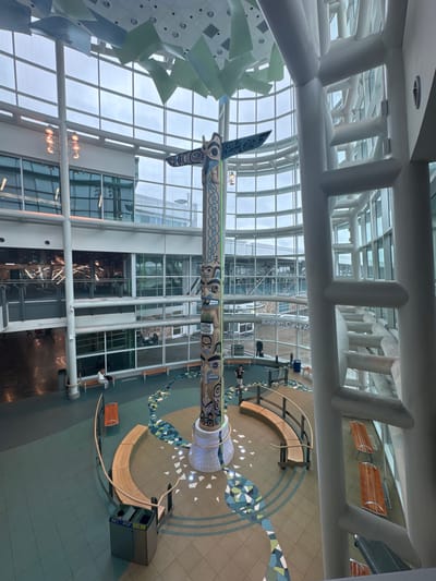 This is a vertically oriented, medium shot taken from an elevated perspective, looking down into a large, modern interior space. The scene appears to be an airport terminal or a similar public building with a high, arched glass ceiling supported by a white, industrial-style framework of beams and columns.

The most prominent subject is a large, intricately carved totem pole positioned in the center of the space, reaching up towards the ceiling. The totem pole features detailed carvings of stylized faces and figures in blues, greens, and blacks, with a long, patterned section extending horizontally from its top.

Around the totem pole, the floor is covered in tiles with a mosaic-like pattern of geometric shapes in green, blue, and white. There are curved wooden benches and seating areas, some with metal supports, and a large grey trash receptacle. A few individuals are visible on the ground level, appearing to be either waiting or walking through the space. One person is seated on a bench, another is standing near the totem pole, and a few others are further in the background. The lighting suggests it is daytime, with natural light filtering through the large glass panes, though the sky outside appears overcast.

In the background, through the glass walls and structural elements, one can see other sections of the building, including staircases and further interior spaces. A sign with white text on a dark background is partially visible on a pillar, reading "Graham Clarke At". The overall atmosphere is one of spaciousness and modern design, with a cultural element introduced by the totem pole.