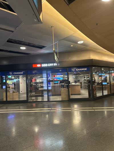 The image depicts the interior of Zurich Airport in Kloten, Switzerland. The camera is positioned at an angle, showing the "CHANGE" office of SBB CFF FFS (Swiss Federal Railways) located in the terminal. The office appears to be a currency exchange service. The flooring is a grey, tiled material with inlaid, parallel, linear lighting. The walls and ceiling have modern, geometric designs with integrated lighting. The store has a clock, with various displays visible through the glass windows. There are no people visible in the immediate frame. The environment indicates a busy transport hub.