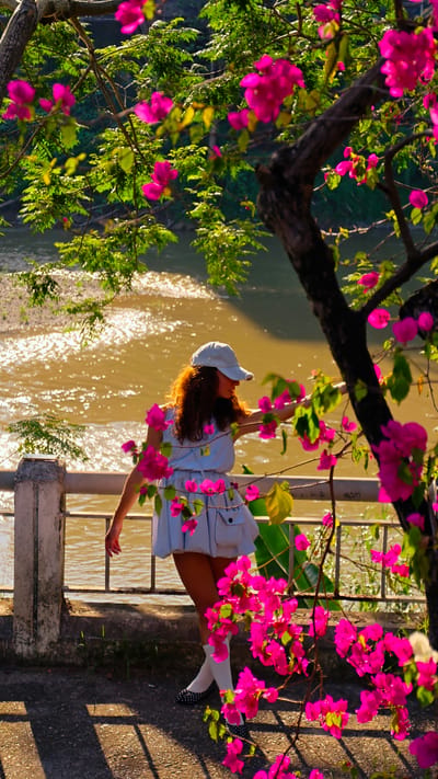 A young woman stands on a stone patio near a railing overlooking a river. She is wearing a light blue dress, white socks, and black and white polka-dotted shoes. Her brown hair is visible under a light-colored baseball cap. She is facing the river and touching the railing with her right hand. Pink bougainvillea flowers frame the scene. The river reflects sunlight, and trees are visible on the opposite bank. The scene is in Lao Cai Ward, Vietnam.