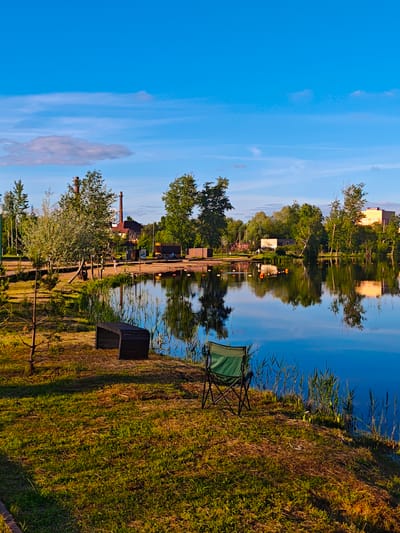 This image depicts a serene waterside landscape in Zaprudnya, Russia, captured during what appears to be the golden hour of either early morning or late afternoon, evidenced by the warm, directional light and elongated shadows on the grass. The sky is a vibrant, clear blue with a few scattered, wispy white clouds.

In the immediate foreground, a lush green and brown grassy bank leads down to calm, reflective water. A dark green folding camping chair is set on the right side of the bank, suggesting a spot for relaxation or observation. To the left, a dark wooden bench is partially obscured by grass and a small tree. The water beautifully mirrors the sky, trees, and distant structures.

On the far bank, a dense line of trees partially screens a collection of buildings. A tall, reddish-brown brick industrial chimney is a prominent feature rising above the tree line, indicating a nearby settlement. Other buildings, some with reddish roofs and others lighter in color, are also visible. Closer to the water's edge on the far side, a sandy or dirt patch is discernible, where two small figures, possibly people, are faintly visible. Several orange buoys are scattered across the water, likely marking a specific area.

The scene conveys a sense of peaceful leisure and natural beauty, subtly blended with hints of human activity and presence. There is no visible text in the image.