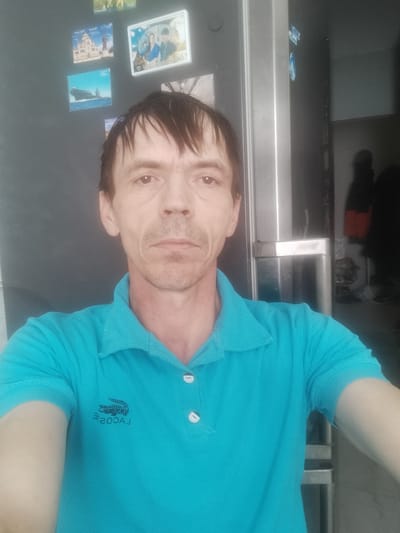 The image is a selfie taken by a man indoors. He is wearing a turquoise polo shirt with a Lacoste logo on the left chest. The man has fair skin, short brown hair, and a thin build. He appears to be in his 40s or 50s. His expression is neutral.
Behind him, on a dark wall, there are several magnetic photos. Some of these appear to be of landmarks and perhaps a couple. To the right of the man, a stainless steel appliance, possibly a refrigerator, is visible. Further in the background, out of focus, there are dimly lit areas and the silhouettes of people. The overall lighting suggests it might be daytime, but the indoor setting makes it difficult to determine. The location context provided is Zyuze, Russia. There is no discernible text in the image except for the brand logo on the shirt, which is partially obscured and appears to be reversed.