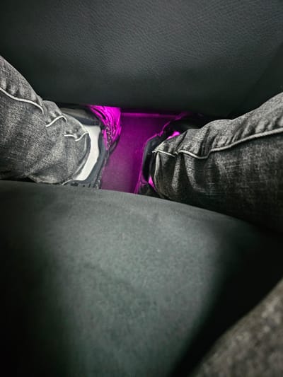 The image shows a close-up view of a person's legs and feet inside a vehicle. The person is wearing dark wash denim jeans and athletic shoes. The shoes appear to be sneakers with white soles and black uppers. Underneath the person's legs, there is a bright pink light emanating from the floor of the vehicle, possibly from ambient lighting or an aftermarket light kit. The interior of the car is dark, with the texture of leather seats visible in the foreground and background. The perspective is low, looking upwards towards the person's legs. There are no visible identifying features of the person or the vehicle's make and model. The location is identified as Montgeron, France.
