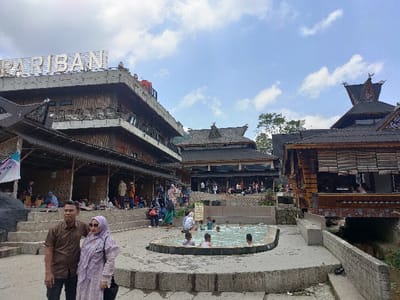 The image depicts a crowded outdoor leisure complex at Pemandian Air Panas Pariban in Semangat Gunung, Indonesia, under a partly cloudy sky. In the immediate foreground, a man in a brown short-sleeved collared shirt and dark pants stands beside a woman wearing a light purple patterned dress, a matching hijab, and sunglasses; both are facing the camera. Behind them, a roughly kidney-shaped hot spring pool contains approximately seven individuals, mostly children, engaging in bathing. The pool is surrounded by concrete terracing and steps.
Dominating the left side of the midground is a multi-story building with a concrete top level displaying the white letters "PARIBAN" and lower levels featuring wooden lattice work and an open-air covered area. Numerous people are visible congregating on the stairs and under the covered sections of this building, some seated on mats. To the right of the pool and further into the background, multiple other buildings with distinct traditional Indonesian architectural elements, including ornate, multi-tiered dark roofs with pointed gables, are visible. One prominent building on the far right features wooden construction and slatted windows. Green foliage and trees are present in the background, indicating a natural setting. The ground consists of concrete pathways, steps, and retaining walls, with some exposed earth or rock.