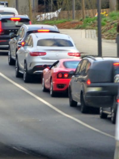 The image displays a queue of vehicles on a paved road in Schifflange, Luxembourg, viewed from a slightly elevated position behind the rearmost visible car. The vehicles are stationary or moving slowly, indicated by multiple illuminated brake lights.
From the foreground right, a portion of a dark-colored SUV is visible, followed directly by a red Ferrari 360 Modena sports car, identifiable by its distinct quad round taillights. Behind the Ferrari is a silver Mercedes-Benz GLE Coupe, bearing "AMG Line" text on its rear bumper and "GLE Coupe" badging on the trunk, with its brake lights illuminated. Further back in the queue, a dark-colored SUV is visible, followed by a dark-colored Volvo SUV, both with illuminated center high-mount stop lamps.
The road surface is dark asphalt, marked by a solid white line defining the right edge of the lane. To the right of the road, a raised curb borders an earthen embankment with sparse deciduous trees, light green vegetation, and small clusters of yellow flowers. A wooden fence runs along the top of the embankment in the background, and a grey street light pole stands on the right side of the scene. No individuals are explicitly visible.
