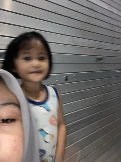 This is a vertically oriented, low-angle, blurry, close-up selfie of two people. The foreground subject is the upper portion of a face, featuring an eye, eyebrow, and forehead with some blemishes, covered by a light gray hood. To the left and slightly behind is the face of a young child with dark hair, dark eyes, and rosy cheeks. The child is wearing a light blue and white tank top with cartoon penguin graphics. The background consists of a corrugated metal wall with vertical ridges. The lighting suggests it is daytime, possibly outdoors, given the natural light. The overall mood appears casual and intimate. No text is visible in the image. The location context is Watampone, Indonesia.