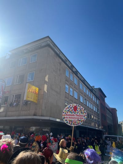 The image shows a building in Köln, Germany, located at coordinates (latitude: 50.926, longitude: 6.958). The building has a tan brick facade with many windows. A sign for the "Kumede Theater" of the "Heimatverein Köln" is held by a person in the image. The sign is a white circle with colorful dots and a red balloon. The sign is held up in front of a crowd of people wearing costumes and masks. The crowd is on the street in front of the building. The sky is blue and clear, and the sun is shining brightly. There is a "Netto" sign on the building. The scene is a street festival, likely part of the annual Cologne Carnival.