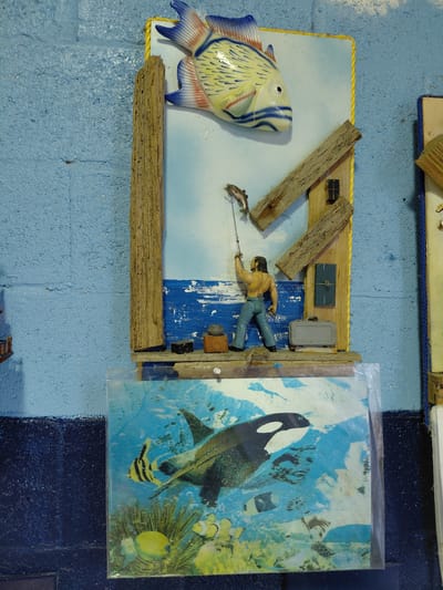 The image shows a diorama or art piece. The scene depicts a fisherman in a small, stylized wooden boat pulling in a fish, with a large ceramic fish figure positioned to the side. The boat and surrounding "water" are constructed from wood, and a small structure resembling a shack or cabin is also included. Next to the boat scene is a framed image of an orca and various other fish. The entire composition is set against a light blue wall, which has a slight texture suggesting it may be concrete or plaster. The lighting is somewhat even, with no strong shadows to suggest a single prominent light source. The location is indicated as Cutler Bay, United States of America.