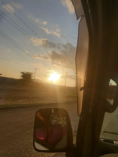 The image presents an outdoor scene at either sunrise or sunset, viewed from inside a vehicle in Mumbuni North ward, Kenya. A bright sun disc is low on the horizon, casting a strong golden light and lens flare across the landscape, which features distant hills or elevated terrain. The sky is light blue with scattered white and grey clouds. Numerous utility poles with overhead wires run parallel to a road on the left side of the frame. Sparse trees and scrub vegetation are visible in the background. In the foreground, a dark, possibly wet, paved road is present, bordered by a dirt or gravel shoulder. The right side of the image shows a portion of the vehicle's dark interior, including a structural pillar and an overhead element. An external side-view mirror, located in the lower-left quadrant, reflects an individual wearing a dark top with a light, intricate pattern and a bright pink or magenta lower garment. No other individuals or specific actions are discernible in the external environment.