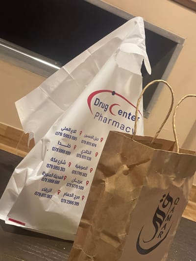 The image captures a close-up, slightly overhead view of two shopping bags placed on what appears to be a flat, dark-colored surface, possibly a table or counter, indoors. Behind the bags, a light beige wall and a dark recess (perhaps a doorway or shelf) are visible.
The prominent item in the foreground is a white plastic bag, partially open and slightly crumpled. The bag features a red and blue oval-shaped logo with the words "Drug Center" in red, sans-serif font, and "Pharmacy" beneath it in a similar, smaller font. A blue dot is visible within the "Drug Center" logo. The white bag is adorned with numerous Arabic location names, each accompanied by a phone number, suggesting various branches of the pharmacy. These locations and their corresponding phone numbers are:
- جبل الحسين (Jabal Al Hussein): 078 8111 874
- تلاع العلي (Tla' Al Ali): 078 5953 595
- الخالدي (Al Khalidi): 078 8111 038
- خلدا (Khalda): 078 5968 699
- الصويفية (Al Sweifieh): 078 5955 393
- شارع مكة (Mecca Street): 0787745 503
- عرجان (Arjan): 078 5770 500
- المدينة المنورة (Al Madinah Al Munawwarah): 078 5968 688
- مرج الحمام (Marj Al Hamam): 078 2966 399
- الزرقاء (Zarqa): 078 8111 063
To the right of the white plastic bag, and slightly behind it, is a brown paper bag with twisted paper handles visible at the top. This bag has a large, stylized logo printed in dark brown or black. The logo appears to be the word "QAMAR" in a flowing, calligraphic script, with its Arabic equivalent "قمر" (Qamar, meaning 'moon') also visible. The paper bag has some creases and wrinkles, indicative of typical paper bag usage.
The lighting in the image suggests an indoor setting, likely with artificial light. The overall scene implies that items have recently been purchased from a pharmacy and another establishment, possibly a food or retail store, and are now resting on a surface.