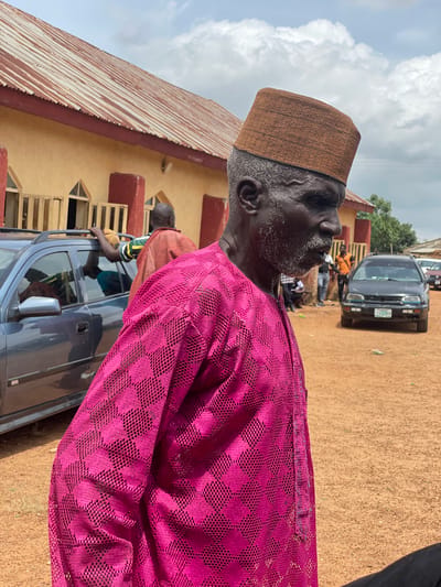 This is a vertical, medium shot of an elderly man in profile, facing left. He wears a brown, textured cap and a vibrant magenta, lace-like fabric garment. His weathered face, with a white beard and mustache, is turned upwards, and he appears to be looking at something in the sky. The background suggests an outdoor gathering in Ban, Nigeria.

Behind the man, a single-story building with a corrugated metal roof and yellow walls is visible. Several people are gathered around a dark gray car, which is parked on a dirt road. One person in an orange shirt is leaning on the car. The ground is covered in reddish-brown dirt. The sky above is partly cloudy with bright white clouds against a pale blue sky. The scene appears to be set during the daytime, possibly late morning or early afternoon, given the brightness of the light. The overall mood is relaxed and informal, perhaps a community event or a social gathering. There is no visible text in the image.