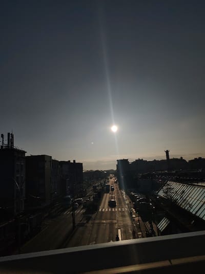 The image captures an urban street scene in Neuilly-Plaisance, France, observed from an elevated vantage point, possibly a bridge or overpass. A bright, circular sun with a prominent vertical lens flare is positioned in the upper center-right, creating strong backlighting that silhouettes most foreground and mid-ground elements. The sky is clear, transitioning from a darker gray at the zenith to a lighter, hazy horizon.
A multi-lane paved road extends from the mid-ground into the distance, with visible white lane markings and pedestrian crosswalks. Numerous vehicles, including cars, vans, and at least one truck, are present on the road, with some appearing in motion. Additional vehicles are parked along the right side of the road.
Multi-story buildings, appearing as dark silhouettes, line both sides of the street. Rooftop communication antennas are visible on structures to the far left. On the right side, a large building features an angled glass roof, and further down the street, a tall, slender tower with a platform near its apex is discernible. Several human figures, rendered as dark silhouettes, are visible on sidewalks and crossing the street. A dark, horizontal railing or ledge occupies the lower foreground, indicating the elevated perspective of the shot.