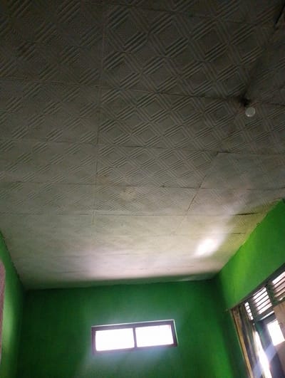 The image shows the interior of a room with green walls and a textured white ceiling. There are two windows visible. One window is in the center of the wall and is rectangular with a lighter frame. The other window is partially visible on the right side of the image and has louvers and curtains. A light fixture is attached to the ceiling. The room appears to be in a residential or commercial building in Sukamanah, Indonesia.