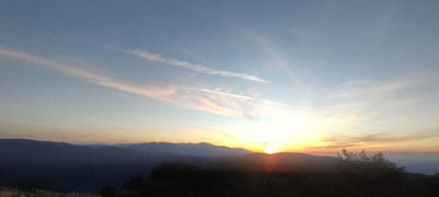 This is a landscape photograph taken at sunrise in Simeonovets, Bulgaria. The scene is dominated by a vast sky with a gradient of light blue at the top transitioning to soft oranges and yellows near the horizon. wispy clouds, some tinged with pink, stretch across the sky, intersected by faint contrails from an aircraft.
Below the sky, a range of silhouetted mountains recedes into the distance, with the closest range appearing dark and indistinct. The foreground is also a dark silhouette of trees and possibly uneven terrain, with some lighter-colored rocks visible at the very bottom left. The sun is partially visible, peeking over the mountain ridge, casting a bright, warm glow that illuminates the lower atmosphere and the edges of the clouds. The overall atmosphere is serene and tranquil, typical of a sunrise in a natural, mountainous environment. No people or specific structures are visible in the image.