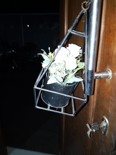 The image shows a black metal plant holder with artificial white flowers hanging from a metal pole attached to a wooden door. A key is inserted into a lock on the door. The background is dark, with some faint light visible in the distance, suggesting it is nighttime or a poorly lit area. The scene appears to be a doorway in Sendangrejo, Indonesia.