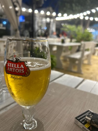 A close-up, eye-level shot focuses on a glass of Stella Artois beer, presented on a light brown, textured tablecloth. The beer is golden yellow with a frothy white head and condensation on the glass. The Stella Artois logo, featuring red and gold lettering, is clearly visible. In the background, a blurry outdoor seating area of a restaurant is depicted, with white tables and chairs, and string lights overhead. The time of day appears to be evening. A partially visible pack of cigarettes and a lighter are also present on the tablecloth. The scene is situated in the city of Aytos, Bulgaria.
