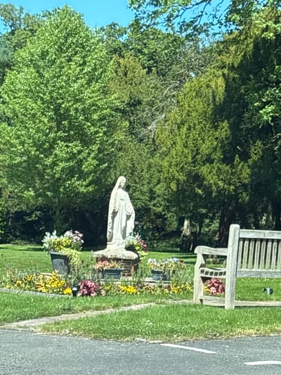 The image shows a section of Wistaston Cemetery in Wistaston, United Kingdom. A white stone statue of a figure, possibly an angel or religious figure, is visible amidst floral arrangements and grave markers. Behind the statue is a wooden bench. The edge of a road is visible on the right, with green grass on either side. Trees form the backdrop. The sky is visible in the upper left.