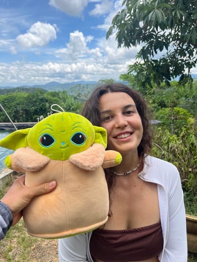 A woman with long, dark curly hair smiles at the camera while holding a plush toy of the character Grogu, also known as Baby Yoda. She is wearing a brown strapless top and an unzipped white jacket over it, along with a beaded necklace. The background features a lush green landscape with rolling hills and a blue sky with white clouds. A body of water is visible to the left of the woman, and trees and foliage surround the area. The scene appears to be outdoors, possibly in a rural or tropical setting in Sơn Điền, Vietnam.