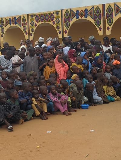A large group of people, mostly children and some adults, are sitting on the ground beside a building with arches and colorful geometric patterns. The ground is dirt or sand. The scene is likely outdoors in Kafanchan A, Nigeria. The people are facing in the same direction, many looking up. Some wear traditional clothing or head coverings.