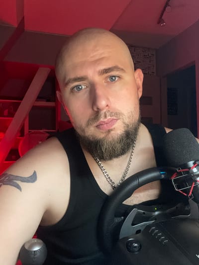 The image shows a close-up, head and shoulders shot of a bald man with blue eyes and a beard. He is wearing a black tank top and a silver chain necklace. He has a tattoo on his left arm. In the foreground, there is a steering wheel and a microphone. The background is dimly lit with red lighting, and appears to be an indoor setting. There are shelves with objects visible in the upper left quadrant of the image. The man is looking directly at the camera. There is no discernible information in the image that can be used to determine the location of the image within the city of Gliwice, Poland.