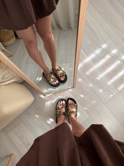A person is standing in a room, looking at their reflection in a full-length mirror. They are wearing a brown dress and leopard print sandals. The room has light-colored wooden flooring and a sheer white curtain in the background. The lighting suggests a sunny day, with shadows from the window falling on the floor. The image is taken from a low angle, focusing on the person's legs and feet. The location is not identifiable as Semiluki, Russia.