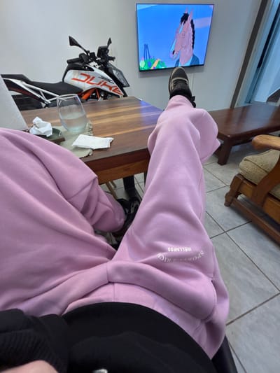 A person is seated, wearing pink sweatpants with the text "SPORTS & RICH WELLNESS" on the upper right leg. They also wear black socks and black and white sneakers. A TV in the background displays a cartoon image of a horse. To the left is a partially visible motorcycle, white with orange and black accents, parked near a wooden table. A glass of liquid and some tissues are on the table. The room appears to be indoors, with a tile floor and wooden furniture. The location is not discernible from the image.