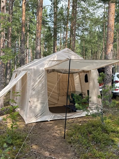 An elevated, eye-level, wide shot captures a large, tan, canvas tent pitched in a forest clearing. The tent has a central open doorway revealing a table and chairs inside. Sunlight filters through the tall, slender pine trees that surround the clearing, casting shadows on the forest floor, which is covered in pine needles, cones, and low-lying green vegetation. A white SUV is partially visible behind the tent to the right, with its rear hatch open. The scene appears to be a campsite, likely in a remote or natural setting.