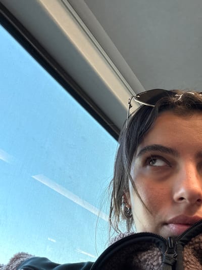 The image depicts a partial close-up of a female individual inside a vehicle, likely a train or bus, in Secaucus, United States. The subject's face is visible from the upper forehead to just below the chin, with her right eye looking upwards and to the left. She has dark hair and wears black "GUCCI" brand sunglasses pushed up onto her head. Two silver-colored hoop earrings are visible in her right earlobe. She is wearing a dark, zipped jacket with a light brown or pinkish-brown textured lining visible around the collar area. The background consists of a large window revealing a clear, bright blue sky with minor reflections or streaks. A dark horizontal frame and a lighter gray upper frame delineate the window structure.