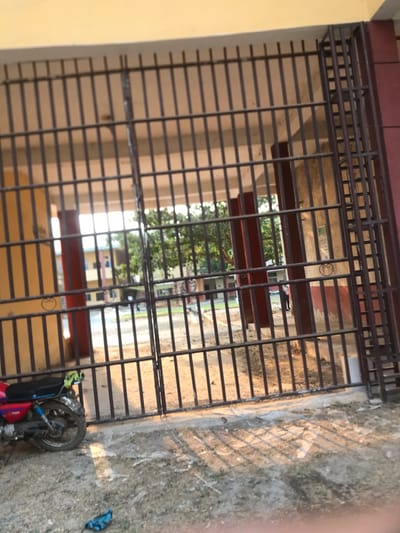 This image captures a ground-level view through a large, dark metal gate, likely an entrance or perimeter barrier, in Umudike, Nigeria. The gate features a grid pattern of vertical and horizontal bars. To the immediate left, a light yellow-colored wall abuts the gate structure, with a darker, reddish-brown band above it.
The foreground ground directly outside the gate, on the right side of the image, is unpaved, appearing dusty, sandy, and somewhat uneven. Strong, elongated shadows from the gate bars are cast across this ground, suggesting direct sunlight and either early morning or late afternoon. A red and black motorcycle is lying on its side on this dusty ground in the bottom right corner, with its front wheel and parts of the engine visible. A small blue object, possibly cloth or plastic, rests on the ground further to the right.
Beyond the gate, green trees with dense foliage are visible, indicating an outdoor, possibly green space. Further in the background, past the trees, light-colored buildings with some darker accents can be seen, hinting at a distant residential or institutional complex. The bright light passing through the gate suggests clear, sunny weather. No people or discernible text are visible in the scene, and no specific activity or event is taking place, presenting a still, observational view.