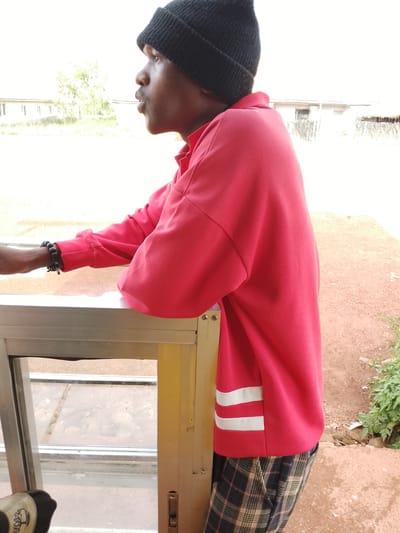 This image features a young Black male standing by a window, looking off to the side. He is wearing a black beanie, a red long-sleeved shirt with two white stripes on the lower left side, and plaid pants. He has a dark bracelet on his left wrist. The setting appears to be outdoors, with a building and vegetation visible in the background. The overall lighting suggests it is daytime, likely a bright day with some overcast conditions or a high sun. The man's expression is neutral, and he seems to be in a relaxed or contemplative state. The window frame is made of metal, and there is a hint of some object in the bottom left corner, possibly a shoe or bag. The ground is a reddish-brown, suggesting an earthy or dusty environment. The location context provided is Agwala, Nigeria.