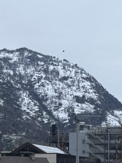 The image depicts a winter landscape in Visp, Switzerland, featuring a snow-covered mountain with sparse, bare trees under an overcast sky. In the foreground, parts of industrial or large commercial buildings are visible. These structures include a building with a corrugated metal roof and a multi-level building featuring external staircases, numerous pipes, ventilation units, and other industrial infrastructure. A light grey street lamp pole with a dark grey fixture extends vertically in the right midground.

In the background, against the upper slopes of the snow-dusted mountain, a small, dark red object, identified as a helicopter, is in flight, positioned to the right of the mountain's crest. Several small, dark structures, possibly houses or religious buildings, are discernible on the mountainside below and to the left of the helicopter. No individuals are visible.